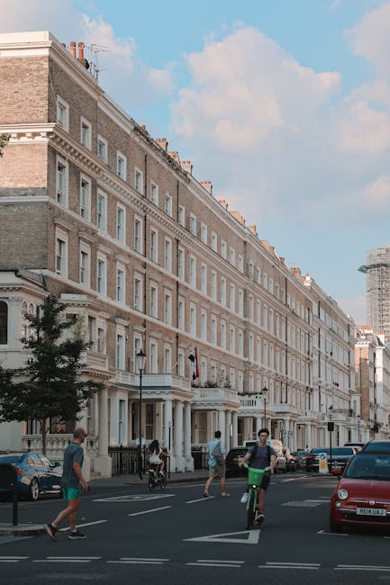 A narrow cobblestone alleyway in a residential area with a gentle curve, lined on the left side by a row of terraced houses featuring white and pastel-colored facades, some with small balconies and wi