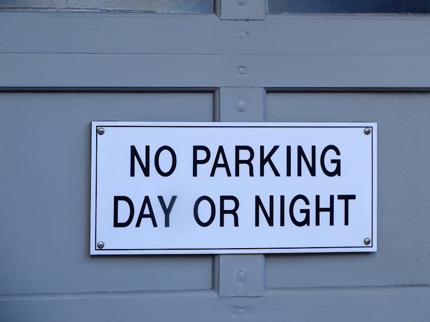 A residential street in W8, Kensington, lined with parked cars on both sides, including compact and mid-sized vehicles in black, silver, blue, and white. The street is shaded by blooming pink cherry b